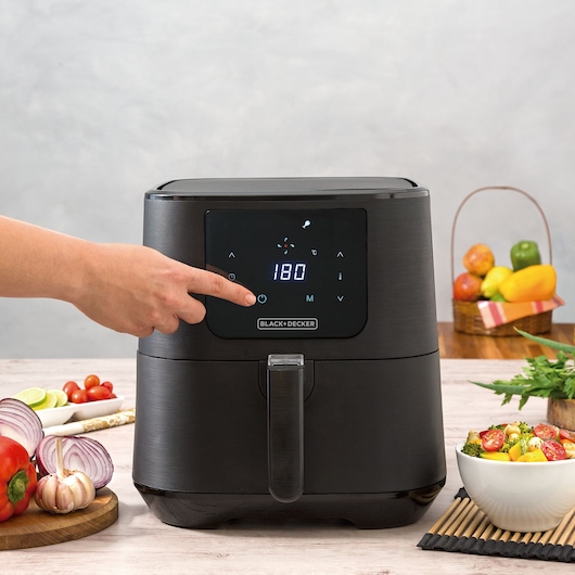 Photo of BLACK+DECKER air fryer ADF7Q with digital display, hand pressing power button, vegetables on table.
