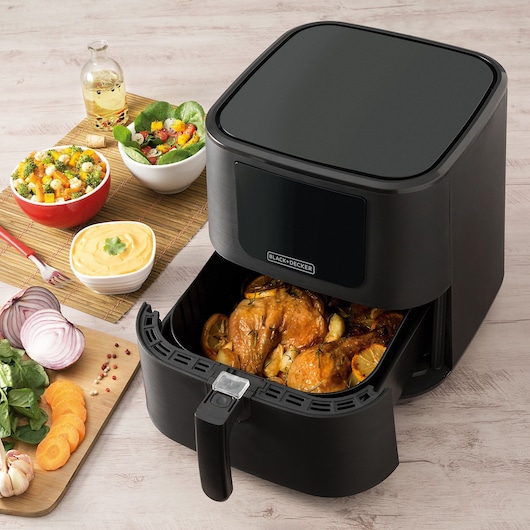 A BLACK+DECKER air fryer is shown on a light wooden table, with its basket open revealing cooked chicken and vegetables inside. Surrounding the air fryer are bowls of salad, a container of sauce, a bottle of oil, and a chopping board with sliced onions, carrots, spinach, garlic, and peppercorns.
