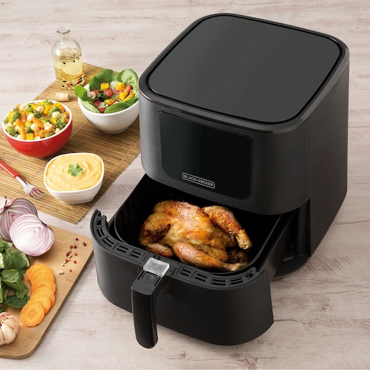 A BLACK+DECKER air fryer with a roasted chicken inside the basket, placed on a light wooden surface. Nearby are bowls of salad and dip, a bottle of oil, and a cutting board with sliced vegetables.