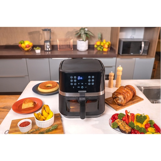 A BLACK+DECKER air fryer is placed on a kitchen counter, surrounded by various types of food including roasted meat, vegetables, fried potatoes, a piece of cake, and a flan. The air fryer has a digital display and touch controls. The kitchen in the background contains fruit bowls, a blender, a microwave, and plants.