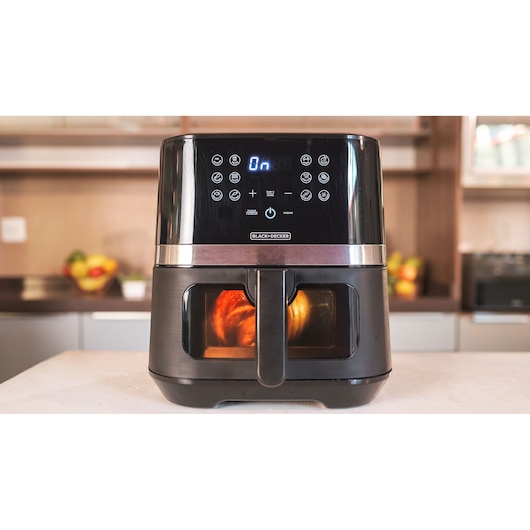 A BLACK+DECKER air fryer with a digital display and control panel is placed on a kitchen counter. The air fryer has a transparent window on the front, showing food being cooked inside.