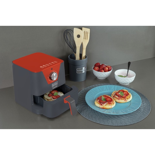 A BLACK+DECKER air fryer with a grey and red design is shown on a kitchen counter. The fryer drawer is open, revealing food inside. Next to it, there is a plate with two small pizzas topped with strawberries and mint leaves. Kitchen utensils in a container, a bowl of strawberries, and a bowl with a spoon are also on the counter.