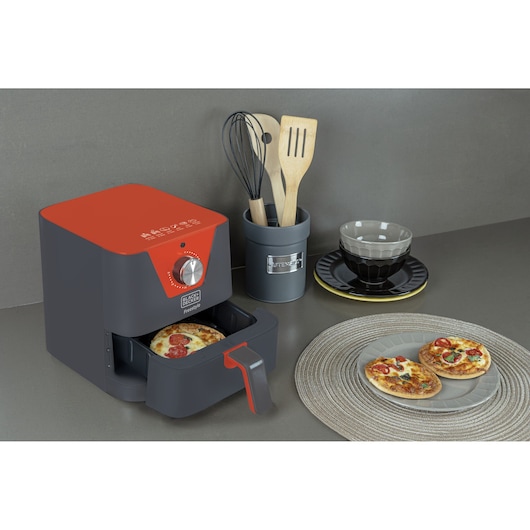 A BLACK+DECKER air fryer with a red top is placed on a kitchen counter. The air fryer drawer is open with a mini pizza inside. Nearby, two mini pizzas are served on a plate on a woven placemat. Kitchen utensils are stored in a container, and a decorative bowl with a saucer is in the background.