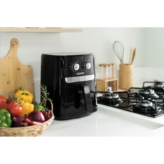 A BLACKDECKER air fryer sits on a kitchen counter next to a gas stove, a basket of colorful vegetables, a wooden cutting board, and kitchen utensils in the background.