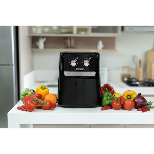 A BLACK+DECKER air fryer is placed on a kitchen counter, surrounded by colorful fresh vegetables such as tomatoes, bell peppers, onion, and chili peppers.
