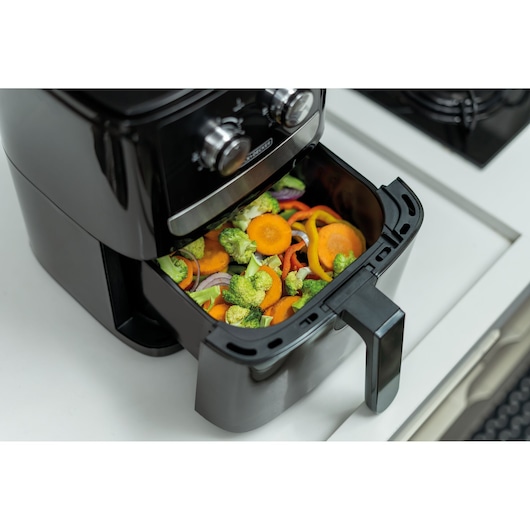 A BLACK+DECKER air fryer filled with sliced fresh vegetables, including carrots, broccoli, onion, and bell peppers, placed on a kitchen countertop.