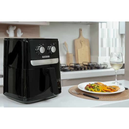 A BLACK+DECKER air fryer sits on a kitchen counter next to a plate of food and a glass of white wine. The air fryer is black with silver dials, and the background shows a modern kitchen with cutting boards and a stove.
