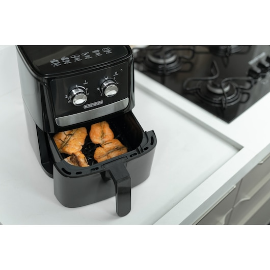 A BLACK+DECKER air fryer with chicken pieces inside, placed on a kitchen countertop next to a gas stove.