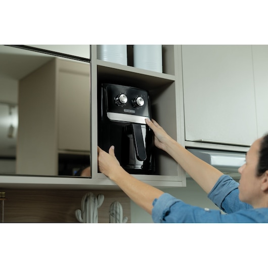 Person placing BLACKDECKER AFM4 black air fryer with two dials into a kitchen cabinet.