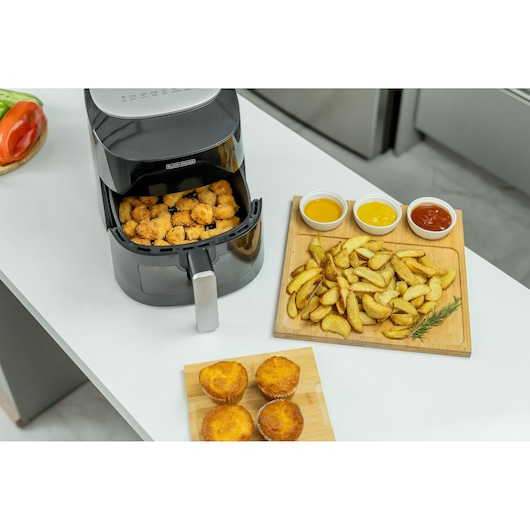 A BLACK+DECKER air fryer on a white kitchen counter with breaded food inside the basket. Next to it is a wooden board with potato wedges and three small bowls of dipping sauces, and a smaller wooden board with four muffins.