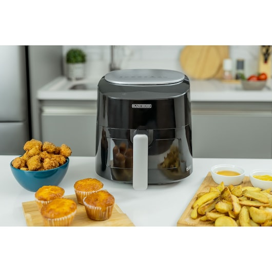 A BLACK+DECKER air fryer is placed on a kitchen counter. Surrounding the air fryer are a bowl of breaded chicken nuggets, four muffins on a wooden tray, a wooden board with potato wedges, and two small bowls with dipping sauces.