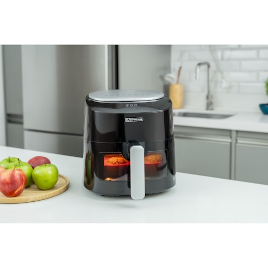 A BLACK+DECKER air fryer with a digital display is placed on a kitchen countertop. There are apples on a round wooden tray next to the air fryer. The kitchen background includes a sink and cabinets.