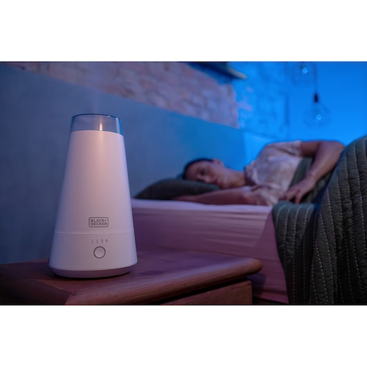 BLACK+DECKER AIR2000N air purifier on bedside table in dimly lit bedroom, person sleeping in background.