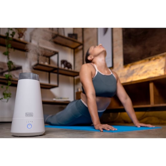 BLACKDECKER AIR2000N humidifier on floor emitting mist, with woman doing yoga in blurred background.