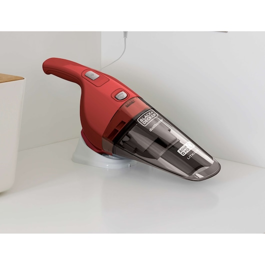 A red BLACK+DECKER handheld vacuum cleaner, model APB3600, is placed on a white charging base on a countertop, designed for versatile cleaning.
