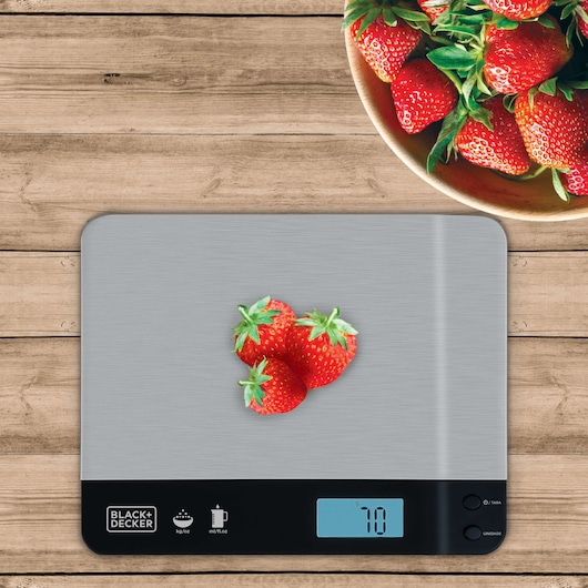 A BLACK+DECKER digital kitchen scale (model BC500) on a wooden surface with three strawberries placed on it and a digital display showing 70 grams. In the top right corner, there is a bowl filled with more strawberries.
