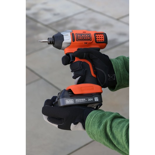 Photo of BLACK+DECKER BDL2020S cordless impact driver, orange and black, held by gloved hands outdoors.