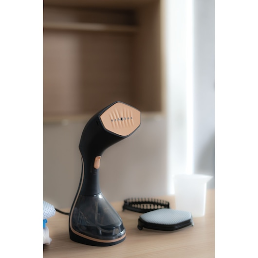 A BLACK+DECKER handheld garment steamer placed on a wooden surface, with attached water tank and accessories including two brush attachments and a measuring cup nearby.
