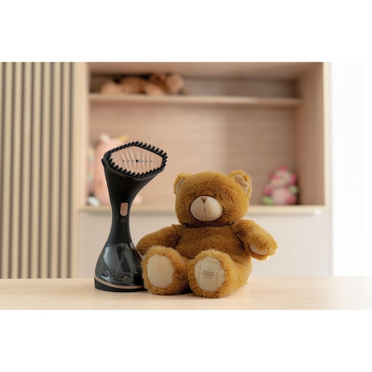 A BLACK+DECKER handheld garment steamer positioned on a wooden table next to a brown teddy bear, with shelves and other toys visible in the background.