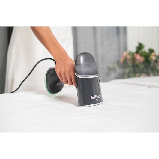A person using a BLACK+DECKER BDV3100 fabric steamer on a white sheet in a home setting.