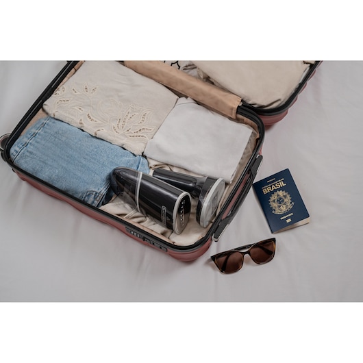 Open suitcase with neatly packed clothes, a BLACK+DECKER handheld vacuum cleaner model BDV3100 inside. Next to the suitcase are a Brazilian passport and a pair of sunglasses on a white surface.