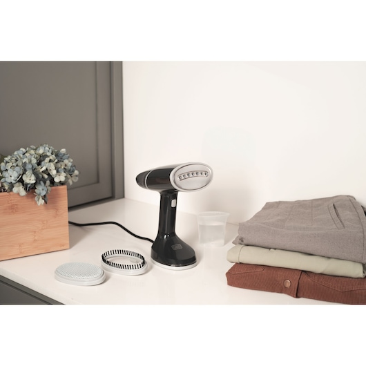 BLACK+DECKER handheld garment steamer with brush attachments beside folded clothes on a countertop.