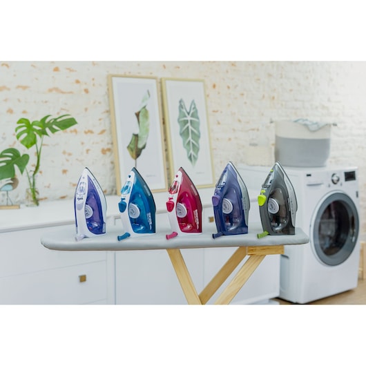 Five colorful BLACK+DECKER steam irons are displayed upright on an ironing board in a modern laundry room, with framed leaf artwork and a washing machine visible in the background.