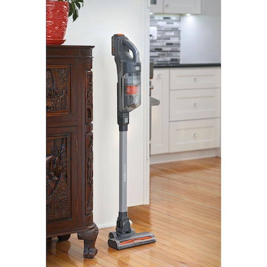 BLACKDECKER BHFEA18D1 cordless stick vacuum cleaner, gray and orange, upright on hardwood floor by cabinet.
