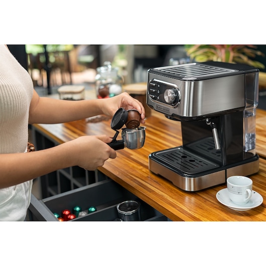Photo of BLACKDECKER CE1100G espresso machine on wooden counter with a person preparing a coffee pod portafilter.