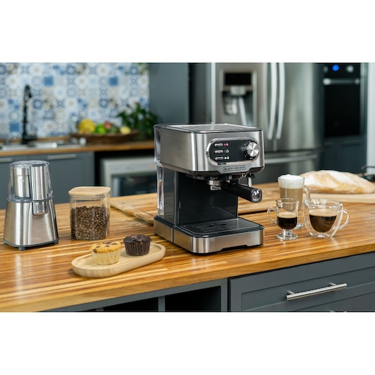 BLACK+DECKER espresso machine set on a kitchen counter, next to a coffee grinder, a jar of coffee beans, two muffins on a wooden tray, and three glass cups of coffee drinks. The kitchen features modern appliances and a decorative tiled backsplash.