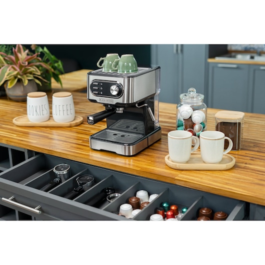 A BLACK+DECKER espresso coffee machine is placed on a wooden kitchen countertop. On top of the machine are two green ceramic cups. Nearby are two white mugs on a light wooden tray, a glass jar filled with coffee pods, a container with ground coffee, and two ceramic jars labeled 'COOKIES' and 'COFFEE'. Below the counter, a drawer is open showing coffee pods and barista tools. The background includes plants and kitchen cabinets.