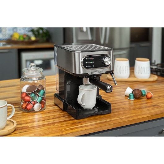 A BLACK+DECKER espresso machine is placed on a wooden kitchen countertop. Nearby, there is a glass jar filled with coffee capsules, a ceramic mug under the machine, and jars labeled 'Coffee' and 'Cookies' in the background. Several coffee capsules are scattered on the counter.