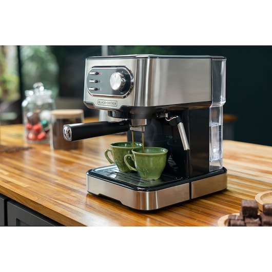 A BLACK+DECKER espresso machine on a wooden kitchen countertop, making coffee into two green cups. Coffee pods and treats are visible in the background.