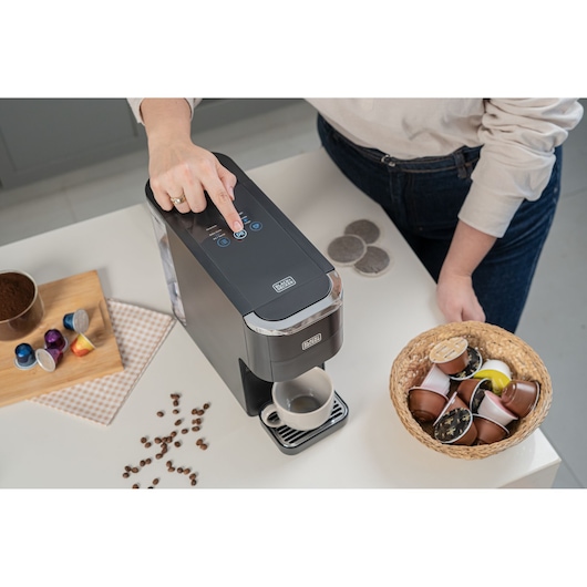 A person is pressing a button on a BLACK+DECKER coffee machine, with a white cup placed below the dispenser. On the table, there are coffee capsules in a basket, coffee pods, coffee beans, and ground coffee, along with several colorful coffee capsules on a wooden board.
