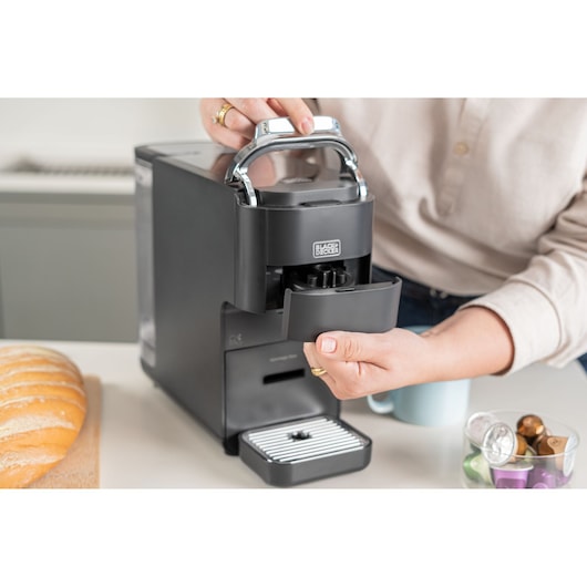 A person operates a BLACK+DECKER coffee machine on a kitchen counter, with a loaf of bread and a bowl containing coffee capsules nearby.