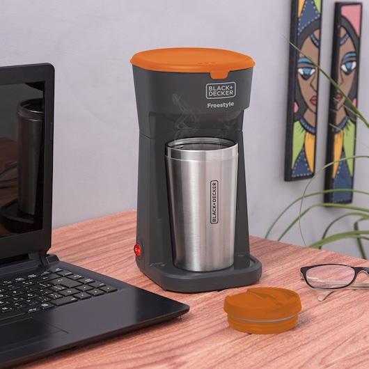 A BLACK+DECKER Freestyle coffee maker with an orange lid and a stainless steel travel mug placed on a wooden desk. The coffee maker is brewing, with steam visible above the mug. A laptop, eyeglasses, and the mug lid are also on the desk. There is a plant and a decorative wall piece in the background.