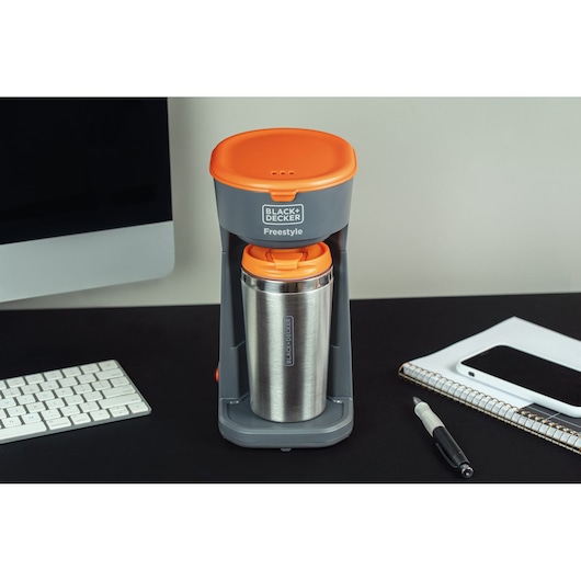 A BLACK+DECKER Freestyle single-serve coffee maker with an orange top and stainless steel travel mug sits on a desk. Nearby are a computer monitor, keyboard, notebook, pen, and smartphone.