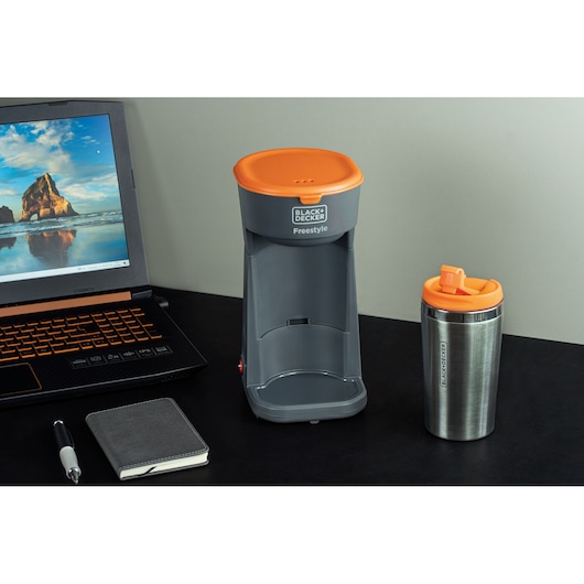 A BLACK+DECKER Freestyle single serve coffee maker in gray with an orange lid is placed on a black desk, next to a matching stainless steel travel mug with an orange lid. Also on the desk are a black laptop, a black notebook, and a pen.