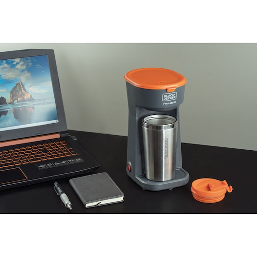 A BLACK+DECKER Freestyle coffee maker with an orange lid is placed on a desk beside a laptop, a pen, a notebook, and the removable orange lid for the coffee maker's stainless steel tumbler.
