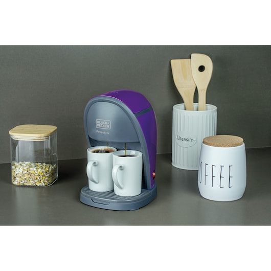 A BLACK+DECKER Freestyle coffee maker in purple and gray is brewing coffee into two white mugs on a kitchen countertop. Next to it are a container with grains, a white jar labeled 'COFFEE' with a cork lid, and a utensil holder with wooden cooking utensils.
