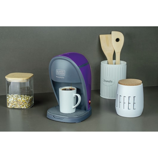 A BLACK+DECKER Freestyle coffee maker in purple and gray is brewing coffee into a white mug. It is placed on a kitchen counter next to a jar with a wooden lid filled with grains, a container labeled 'Utensils' holding wooden kitchen tools, and a ceramic jar labeled 'COFFEE' with a cork lid.