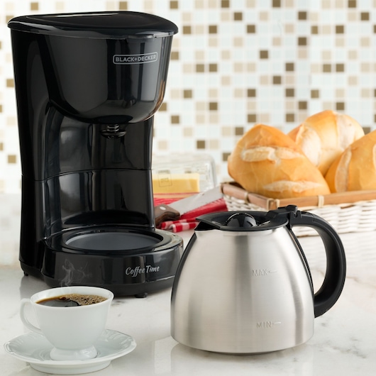 A BLACK+DECKER coffee maker with a stainless steel carafe is displayed on a kitchen counter. In the foreground, there is a cup of coffee on a saucer, and in the background, a basket of bread rolls is visible.