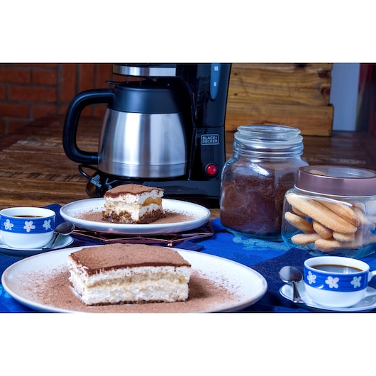 A BLACK+DECKER coffee maker sits on a wooden table next to a jar of ground coffee, a jar of ladyfinger biscuits, two plates with slices of tiramisu dessert, and two cups of coffee in blue and white floral cups with spoons.