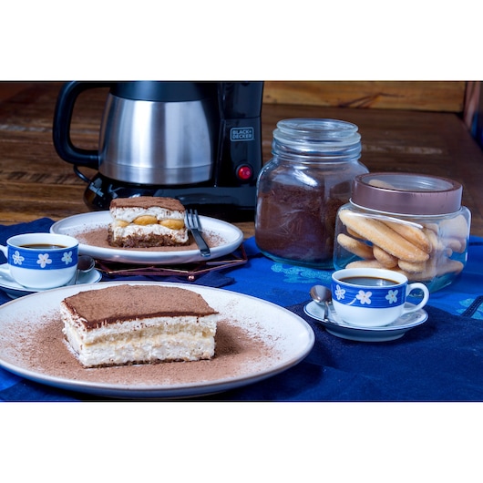 A Black+Decker coffee maker is on a wooden table next to jars of coffee powder and ladyfinger biscuits. Two plates with slices of tiramisu sit in front, accompanied by cups of coffee in blue and white floral-patterned cups and saucers.