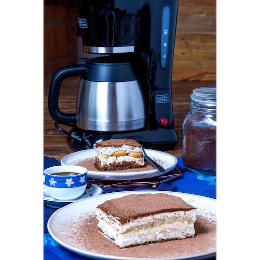 A BLACK+DECKER drip coffee maker on a table next to a glass jar containing ground coffee. In the foreground, there are two plates with pieces of tiramisu dessert, and a cup of coffee. The scene is set on a blue tablecloth with a rustic wooden background.