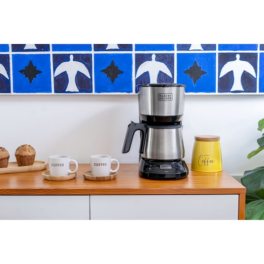 A BLACK+DECKER coffee maker sits on a wooden counter alongside two coffee mugs, a yellow coffee container, and two muffins on a tray. A blue and white decorative painting is visible in the background.
