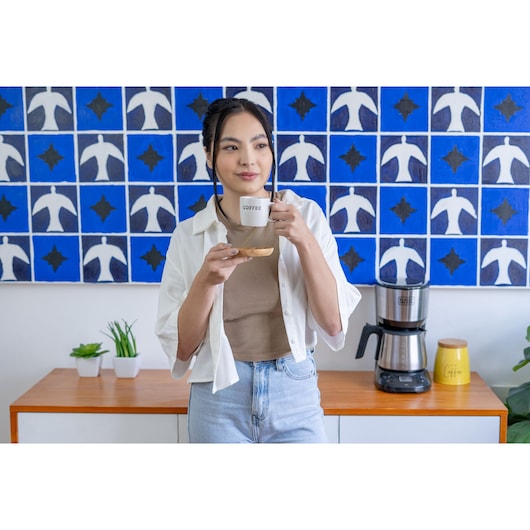 A person standing in a modern kitchen area holding a coffee cup and saucer. On the wooden counter behind them is a BLACK+DECKER coffee maker (model CM250P) next to a yellow jar labeled 'Coffee' and small potted plants. A blue and white patterned artwork hangs on the wall above.