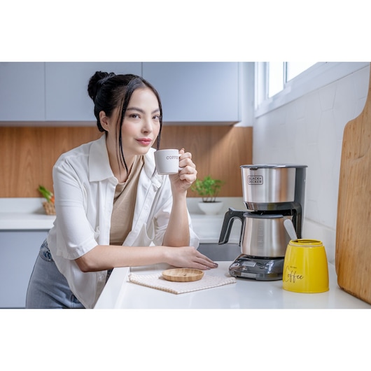 A person in a kitchen holding a small coffee cup near a BLACK+DECKER coffee maker (model CM250P) on a countertop. There is a yellow container labeled 'coffee' and a wooden tray next to the coffee maker.