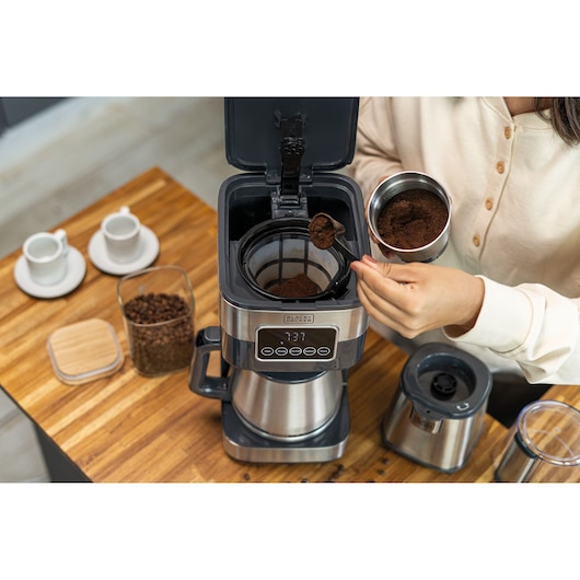 BLACKDECKER CM350G coffee maker front angle with person adding ground coffee, coffee grinder and cups on wooden table.