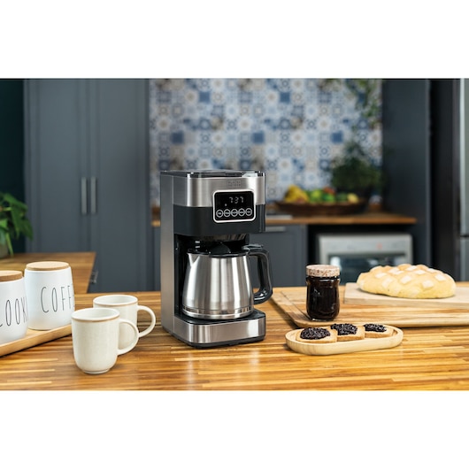 A BLACK+DECKER CM350G coffee maker sits on a wooden kitchen countertop next to coffee mugs, containers labeled 'COFFEE' and 'LOOK', a jar of jam, bread, and toast with jam. The coffee maker has a stainless steel carafe and digital display, with a modern kitchen in the background.